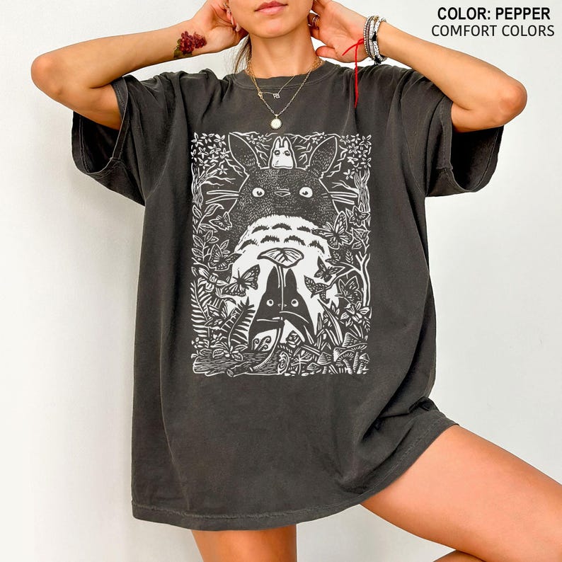 Puede incluir: Camiseta de color pimienta de gran tama&ntilde;o con un gr&aacute;fico blanco de una escena de bosque con una criatura grande, similar a un dibujo animado, y figuras m&aacute;s peque&ntilde;as. El texto "COLOR: PEPPER COMFORT COLORS" es visible.