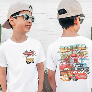 May include: White t-shirt with a cartoon car design. The front displays a yellow car with the word "Name" above it. The back of the shirt features a colourful graphic of various cars and the words "Radiator Springs".