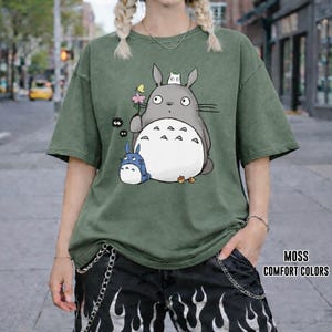 May include: A moss green t-shirt featuring a graphic of a large gray cartoon character with a white belly, holding a flower, with smaller blue and white characters. The shirt has short sleeves and a relaxed fit. The text "MOSS COMFORT COLORS" is visible.