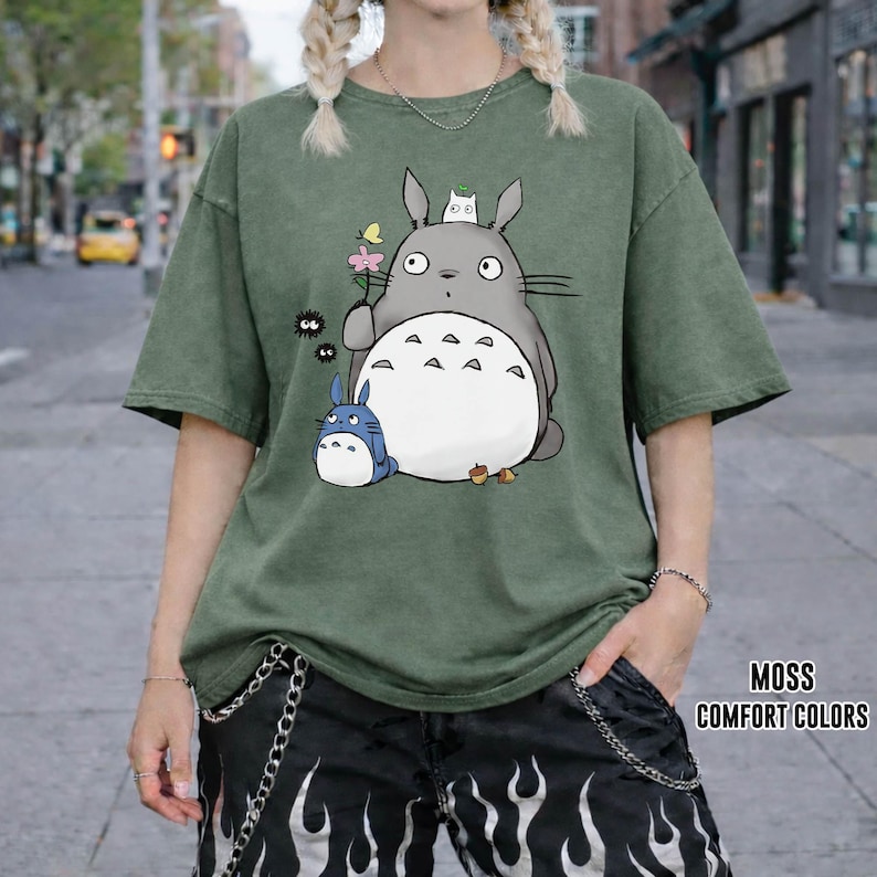 May include: A moss green t-shirt featuring a graphic of a large gray cartoon character with a white belly, holding a flower, with smaller blue and white characters. The shirt has short sleeves and a relaxed fit. The text "MOSS COMFORT COLORS" is visible.