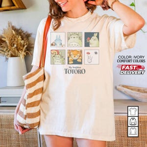 May include: Ivory-colored t-shirt featuring a grid of six cartoon images of characters from "My Neighbor Totoro." The shirt includes the text "My Neighbor Totoro" in a bold font. The shirt also has the text "COLOR: IVORY COMFORT COLORS" and "FAST DELIVERY."