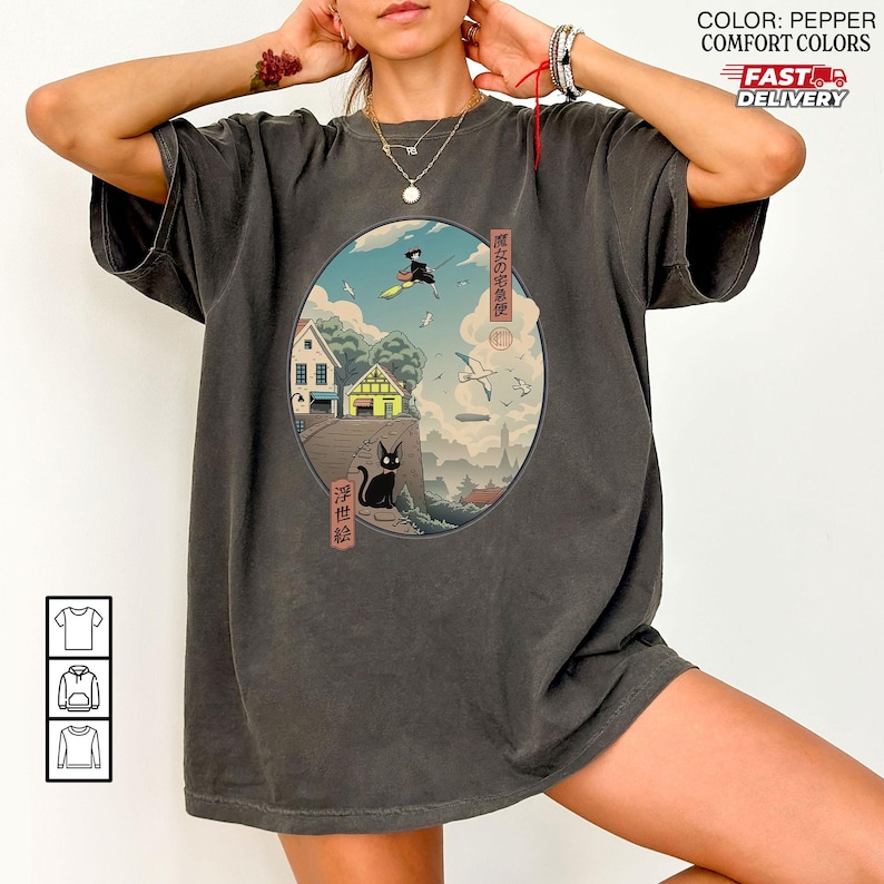 May include: Pepper-colored t-shirt featuring a circular graphic with Japanese art. The graphic depicts a witch flying on a broom, a black cat, and traditional Japanese buildings. The text on the shirt is in Japanese.