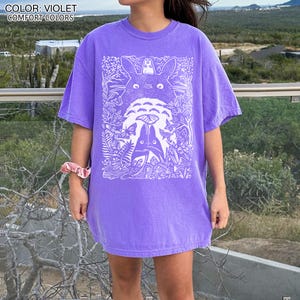 Puede incluir: Camiseta violeta de colores c&oacute;modos con un gr&aacute;fico blanco de personajes de dibujos animados en una escena de bosque. La camiseta tiene mangas cortas y un corte relajado. El texto "COLOR: VIOLET COMFORT COLORS" est&aacute; en la parte superior de la imagen.