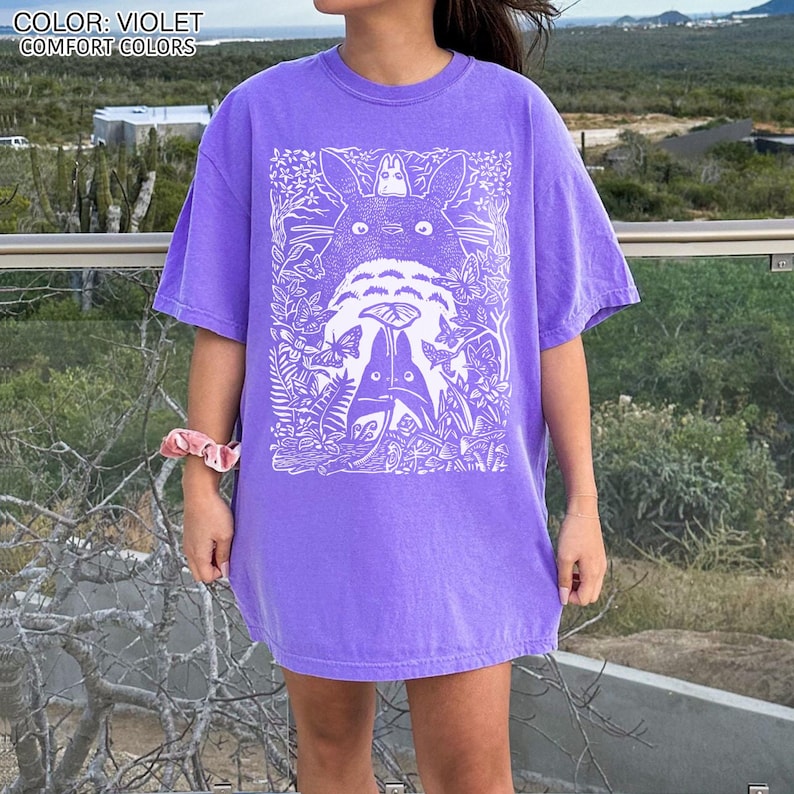 Puede incluir: Camiseta violeta de colores c&oacute;modos con un gr&aacute;fico blanco de personajes de dibujos animados en una escena de bosque. La camiseta tiene mangas cortas y un corte relajado. El texto "COLOR: VIOLET COMFORT COLORS" est&aacute; en la parte superior de la imagen.