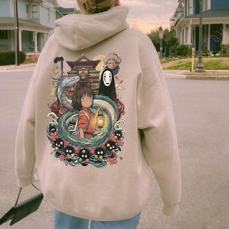 May include: Beige hoodie with a Japanese animated film graphic. The design features a girl with a lantern, a dragon, and other characters. The hoodie has a drawstring hood and long sleeves.