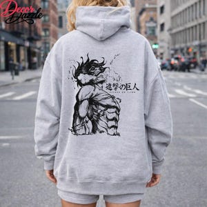 May include: A light gray hoodie featuring a black and white illustration of a muscular figure with the text "Attack on Titan" in Japanese. The hoodie has a hood and long sleeves.