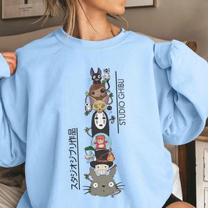 May include: Light blue sweatshirt featuring a stacked graphic of Studio Ghibli characters, including Totoro, No-Face, and Jiji. The words "STUDIO GHIBLI" and Japanese text are printed vertically on the right and left sides of the graphic.