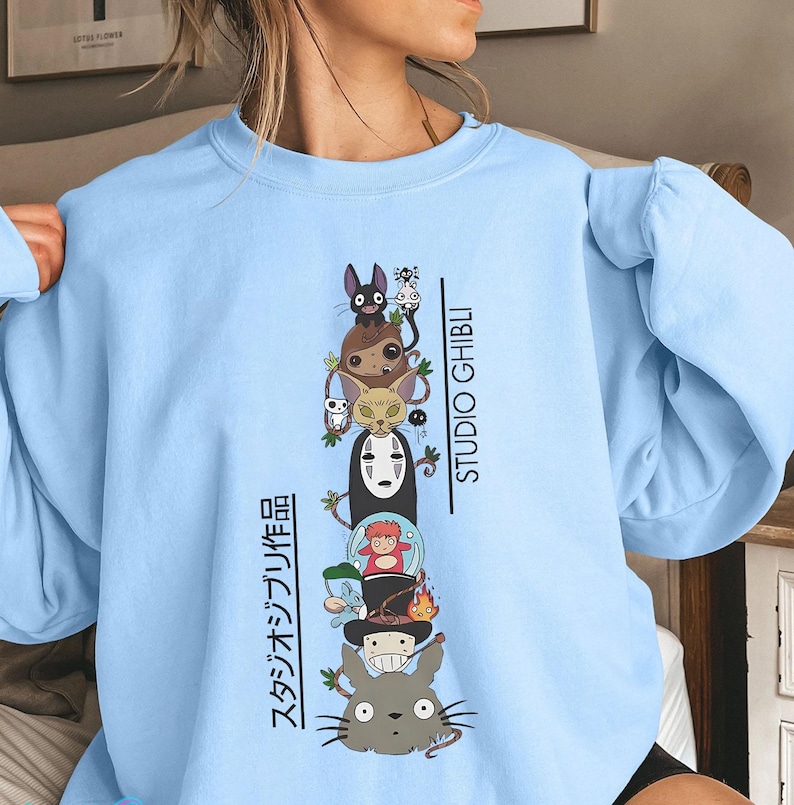 May include: Light blue sweatshirt featuring a stacked graphic of Studio Ghibli characters, including Totoro, No-Face, and Jiji. The words "STUDIO GHIBLI" and Japanese text are printed vertically on the right and left sides of the graphic.