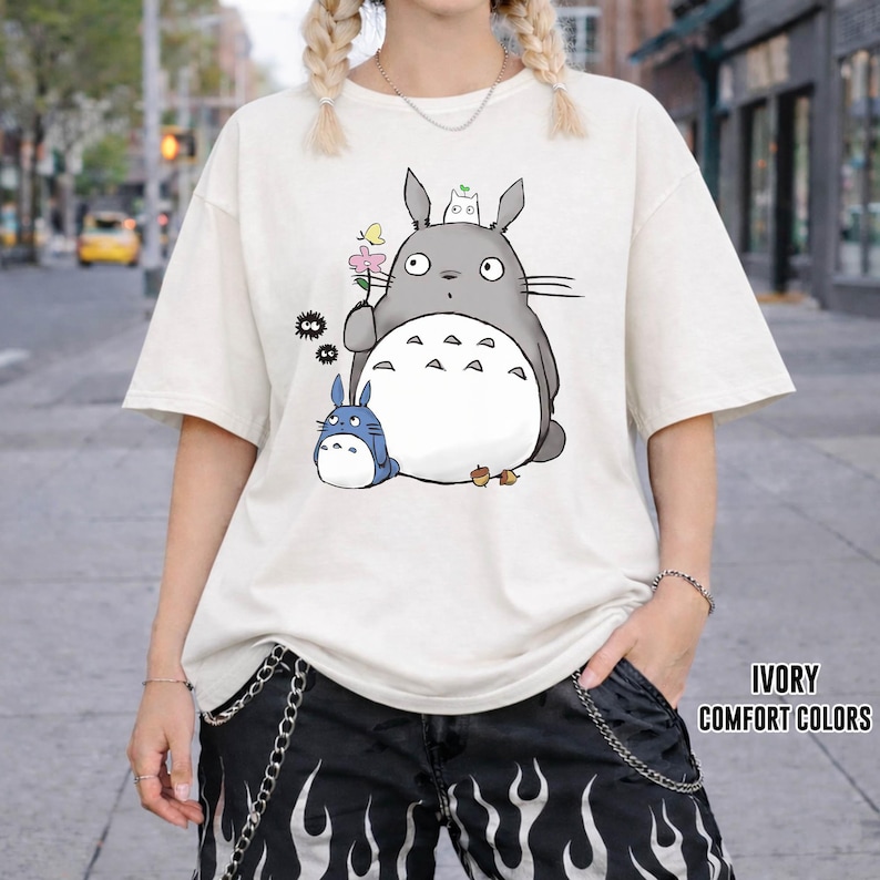 May include: Ivory-colored, oversized t-shirt featuring a graphic of a gray Totoro character holding a flower, with a smaller blue Totoro. The shirt has short sleeves and the text "IVORY COMFORT COLORS".