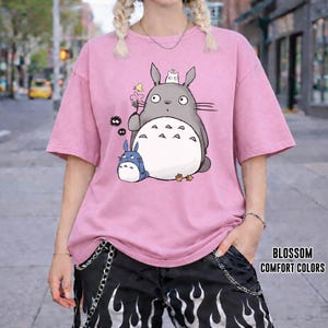 May include: Pink t-shirt featuring a cartoon Totoro design with a small blue Totoro and other characters. The shirt has short sleeves and a relaxed fit. The text "BLOSSOM COMFORT COLORS" is printed on the lower right.