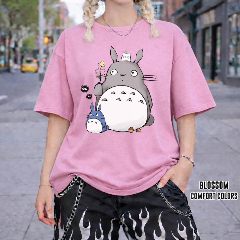 May include: Pink t-shirt featuring a cartoon Totoro design with a small blue Totoro and other characters. The shirt has short sleeves and a relaxed fit. The text "BLOSSOM COMFORT COLORS" is printed on the lower right.