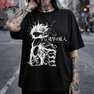 May include: Black t-shirt featuring a white graphic of a muscular figure with the Japanese text "Attack on Titan." The design includes detailed shading and dynamic lines, creating a bold visual statement. The shirt is a classic crew neck style.
