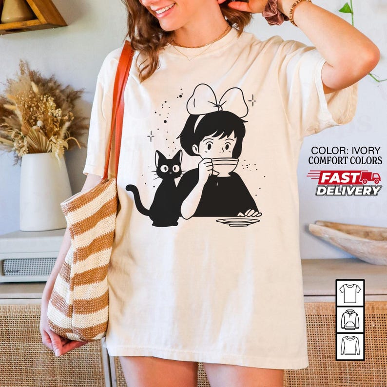 以下が含まれることがあります： アイボリー色のTシャツ。黒と白のイラストで、女の子がカップから飲み物を飲んでおり、黒猫がそばにいます。Tシャツには「COLOR: IVORY COMFORT COLORS」と「FAST DELIVERY」の文字が入っています。