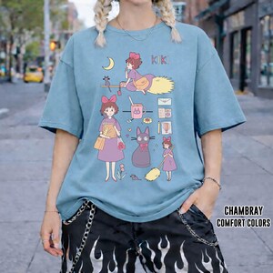 Peut inclure: T-shirt bleu clair avec une illustration de dessin anim&eacute; avec le mot "KIKI". Le motif comprend une sorci&egrave;re sur un balai, un chat noir et d'autres graphiques connexes. Le t-shirt est en mati&egrave;re souple.