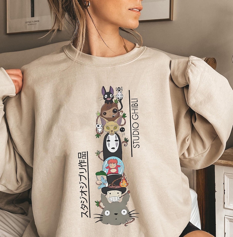 May include: Beige sweatshirt featuring a stacked graphic of Studio Ghibli characters, including Totoro and No-Face. The design includes the text "STUDIO GHIBLI" and Japanese text. The sweatshirt is a neutral color.