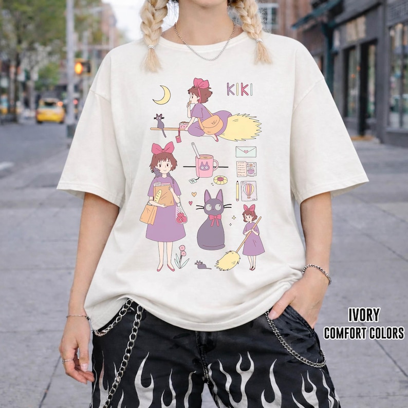 Peut inclure: T-shirt ivoire avec un motif de dessin anim&eacute; repr&eacute;sentant une jeune fille, un chat noir et le mot "KIKI". Le motif comprend un croissant de lune, un balai et d'autres illustrations. Le texte "IVORY COMFORT COLORS" est imprim&eacute; en bas &agrave; droite.