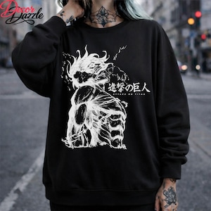 May include: Black sweatshirt featuring a white graphic of a muscular figure with spiky hair and the Japanese text "Attack on Titan." The design is a detailed illustration. The sweatshirt is a casual garment.