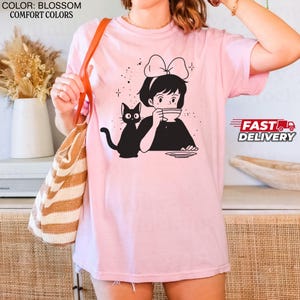 以下が含まれることがあります： 女の子がカップから飲み物を飲んでいる白黒イラストが描かれた、明るいピンク色のTシャツ。横には黒猫がいます。Tシャツの右側には「FAST DELIVERY」の文字があります。
