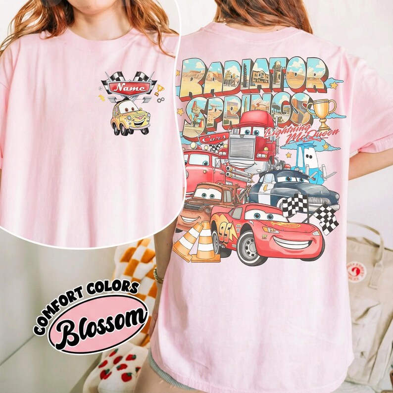 May include: Pale pink t-shirt with a cartoon car design on the front, including the word "Name". The back showcases a vibrant graphic of cartoon cars and the text "Radiator Springs" and "Lightning McQueen".