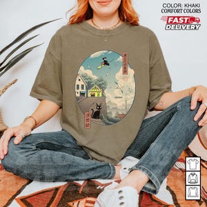 May include: Khaki-colored t-shirt featuring a circular graphic with Japanese art. The graphic depicts a witch on a broom, a black cat, houses, and birds. The text "COLOR: KHAKI COMFORT COLORS" and "FAST DELIVERY" are also visible.