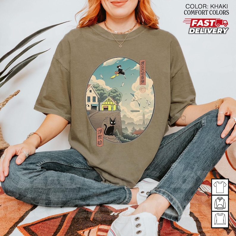 May include: Khaki-colored t-shirt featuring a circular graphic with Japanese art. The graphic depicts a witch on a broom, a black cat, houses, and birds. The text "COLOR: KHAKI COMFORT COLORS" and "FAST DELIVERY" are also visible.