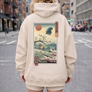May include: Beige hoodie featuring a Japanese anime-style illustration on the back. The artwork depicts a landscape with figures, a red sun, and Japanese text. The hoodie has a hood and a relaxed fit.