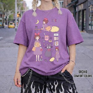 Peut inclure: T-shirt violet avec un dessin anim&eacute; d'une jeune fille sur un balai, un chat noir et d'autres illustrations. Le mot "KIKI" est imprim&eacute; au-dessus du dessin. Fait partie de la collection "ORCHID COMFORT COLORS".