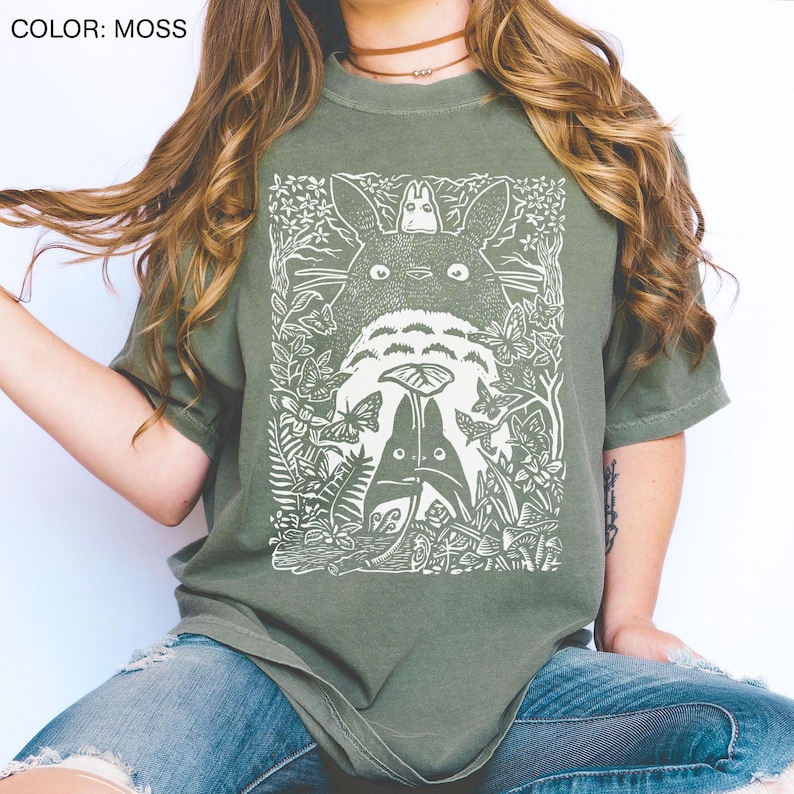 Puede incluir: Camiseta verde musgo con un gr&aacute;fico blanco de una escena de bosque y personajes. El dise&ntilde;o incluye una criatura grande de ojos redondos, figuras m&aacute;s peque&ntilde;as, mariposas y follaje. La camiseta es de estilo casual y c&oacute;modo.