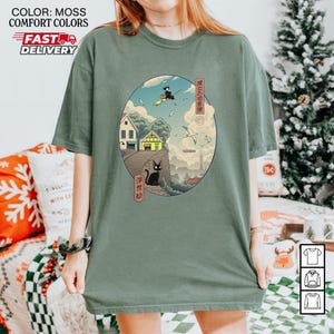 May include: Moss green t-shirt featuring an oval graphic with a Japanese-inspired illustration. The artwork depicts a black cat, a witch on a broom, houses, and text in Japanese. The shirt has the words "COLOR: MOSS COMFORT COLORS FAST DELIVERY" printed on the top left.
