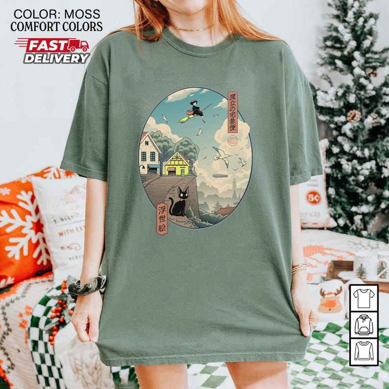 May include: Moss green t-shirt featuring an oval graphic with a Japanese-inspired illustration. The artwork depicts a black cat, a witch on a broom, houses, and text in Japanese. The shirt has the words "COLOR: MOSS COMFORT COLORS FAST DELIVERY" printed on the top left.