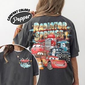 May include: Dark grey t-shirt with a colourful graphic of cartoon cars and the text "Radiator Springs" on the back. The front of the shirt has a small car graphic with the text "Name". The shirt also has the text "Comfort Colors Pepper".