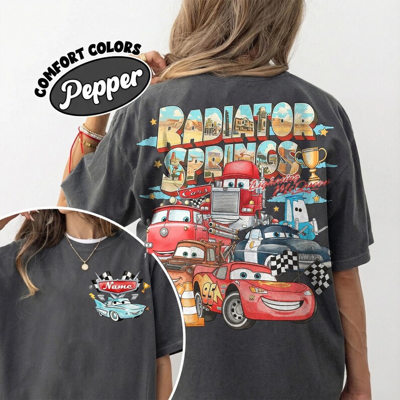 May include: Dark grey t-shirt with a colourful graphic of cartoon cars and the text "Radiator Springs" on the back. The front of the shirt has a small car graphic with the text "Name". The shirt also has the text "Comfort Colors Pepper".