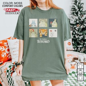 May include: Moss green t-shirt featuring a grid of six colorful illustrations of characters from "My Neighbor Totoro." The text "My Neighbor Totoro" is printed below the illustrations. The shirt has a relaxed fit.