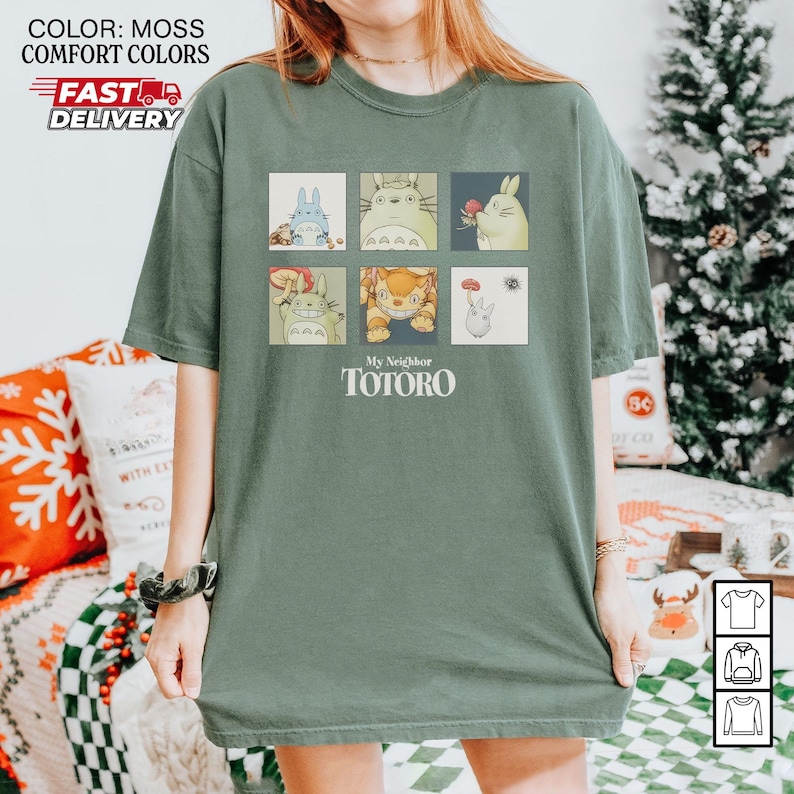 May include: Moss green t-shirt featuring a grid of six colorful illustrations of characters from "My Neighbor Totoro." The text "My Neighbor Totoro" is printed below the illustrations. The shirt has a relaxed fit.