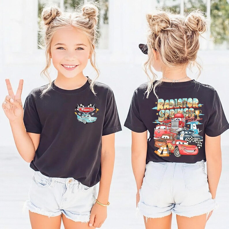 May include: Black t-shirt with a cartoon car graphic on the front and back. The front graphic includes the word "Name" and a light blue car. The back graphic says "Radiator Springs" with various cartoon cars. The shirt is worn with light blue denim shorts.