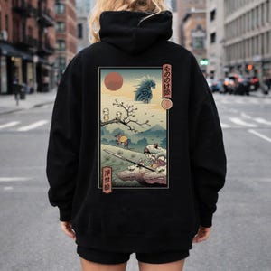 May include: Black hoodie featuring a Japanese anime-style illustration on the back. The artwork depicts a scene with characters, a red sun, and Japanese text. The hoodie has a drawstring hood and a kangaroo pocket.