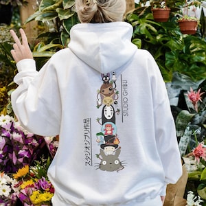 May include: White hoodie featuring a stacked graphic of Studio Ghibli characters, including Totoro and No-Face. The back of the hoodie displays the text "STUDIO GHIBLI" and Japanese text. A casual, comfortable garment.