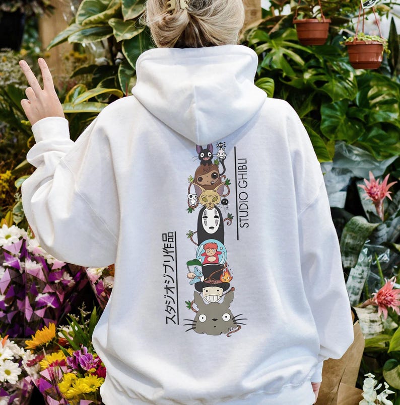 May include: White hoodie featuring a stacked graphic of Studio Ghibli characters, including Totoro and No-Face. The back of the hoodie displays the text "STUDIO GHIBLI" and Japanese text. A casual, comfortable garment.