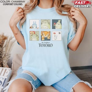 May include: Light blue t-shirt featuring six square illustrations of characters from "My Neighbor Totoro." The shirt includes the text "My Neighbor Totoro" in a stylized font. The shirt is a chambray color and made of a comfortable material.