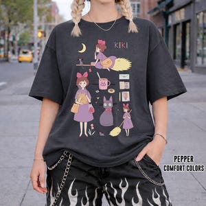 Peut inclure: T-shirt gris fonc&eacute; avec un motif de Kiki la petite sorci&egrave;re. Le design comprend Kiki sur un balai, un chat noir et d'autres illustrations. Le texte "KIKI" est imprim&eacute; en haut. Fait partie de la collection Pepper Comfort Colors.