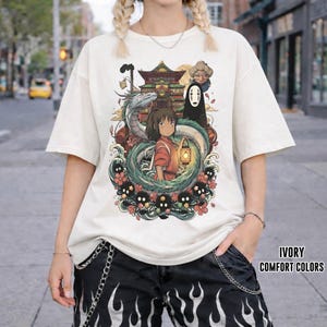 May include: Ivory t-shirt with a vibrant graphic from the animated film Spirited Away. The design showcases characters such as Chihiro, No-Face, and Haku, set amidst floral and dragon elements. The text "IVORY COMFORT COLORS" is printed on the bottom right.