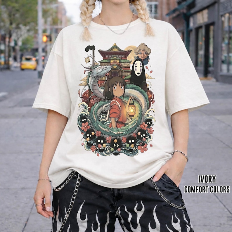 May include: Ivory t-shirt with a vibrant graphic from the animated film Spirited Away. The design showcases characters such as Chihiro, No-Face, and Haku, set amidst floral and dragon elements. The text "IVORY COMFORT COLORS" is printed on the bottom right.