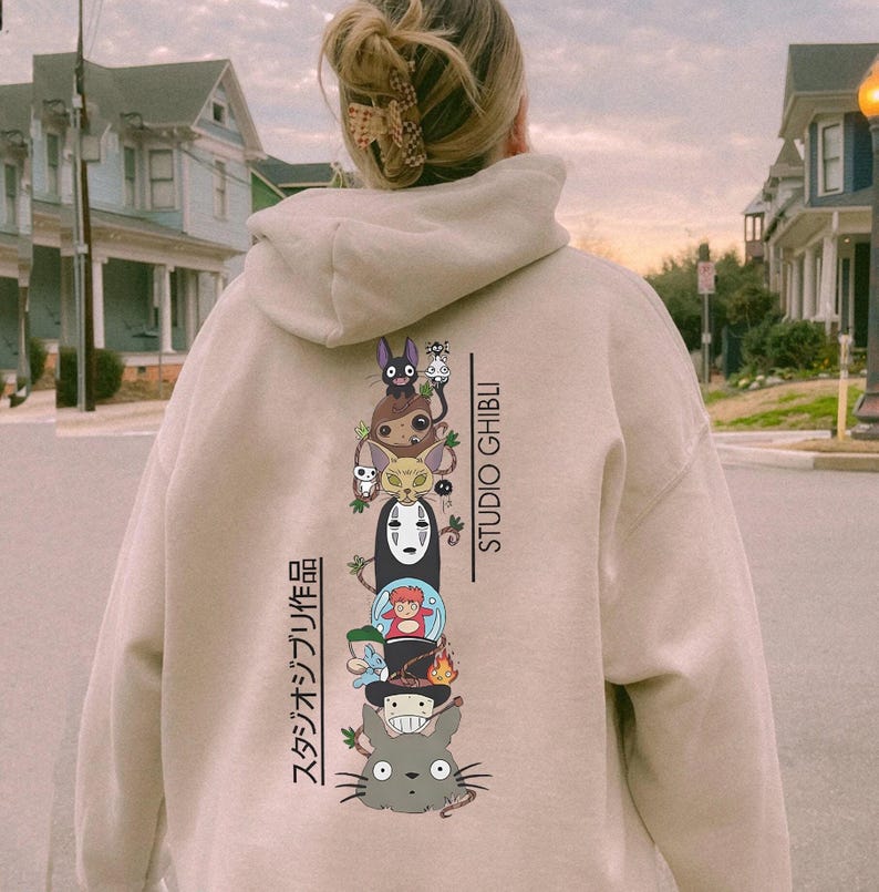May include: Beige hoodie featuring a stacked graphic of Studio Ghibli characters, including Totoro and No-Face. The text "STUDIO GHIBLI" is printed vertically on the right side, with Japanese text on the left. The hoodie has a drawstring hood and a kangaroo pocket.