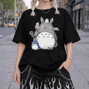 May include: Black t-shirt featuring a cartoon illustration of a large gray creature with a white belly, holding a flower, and a smaller blue creature. The design includes additional small white and black details.