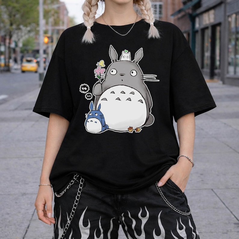 May include: Black t-shirt featuring a cartoon illustration of a large gray creature with a white belly, holding a flower, and a smaller blue creature. The design includes additional small white and black details.