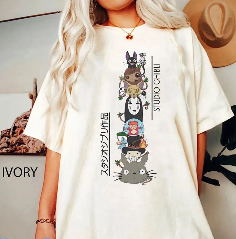 May include: Ivory-colored t-shirt featuring a stacked graphic of Studio Ghibli characters, including Totoro, No-Face, and others. The text "STUDIO GHIBLI" and Japanese text are printed vertically on the right and left sides of the graphic.