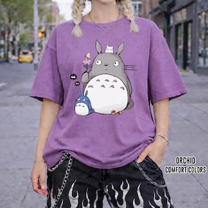 May include: A purple t-shirt featuring a cartoon Totoro design with a small blue Totoro and other characters. The shirt has short sleeves and a relaxed fit. The text "ORCHID COMFORT COLORS" is printed on the bottom right.