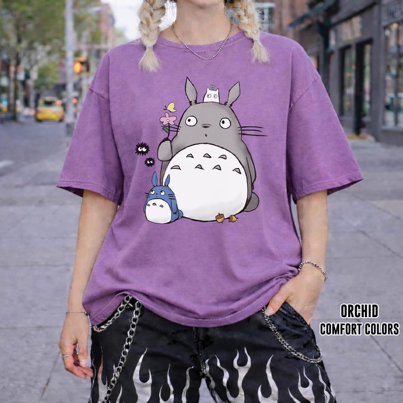 May include: A purple t-shirt featuring a cartoon Totoro design with a small blue Totoro and other characters. The shirt has short sleeves and a relaxed fit. The text "ORCHID COMFORT COLORS" is printed on the bottom right.