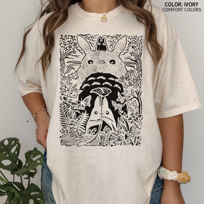 Puede incluir: Camiseta de color marfil con un gr&aacute;fico en blanco y negro de una escena de bosque con tres personajes de dibujos animados. El texto "COLOR: IVORY COMFORT COLORS" est&aacute; impreso en la parte superior derecha. La camiseta tiene mangas cortas y un corte holgado.