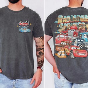 May include: Dark grey t-shirt with a front and back design. The front features a blue car graphic with the word "Name" and racing flags. The back displays a colourful graphic of cartoon cars with the words "Radiator Springs".